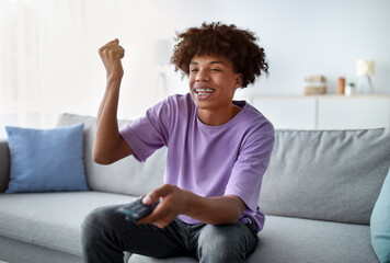 Excited African American teen with remote control enjoying movie, making YES gesture at home. Emotional black adolescent watching TV on couch in living room, cheering, feeling overjoyed