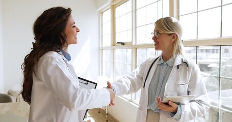 Two women therapists in uniforms lead professional or personal conversation standing together in modern clinic office, smiling, exchange handshake, gesture of mutual respect, collaboration and trust - Powered by Adobe