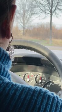 Closeup hands on steering wheel driving, woman in blue sweater with rings steering through city, dashboard and passing roadside visible. Vertical shot taken on mobile device.