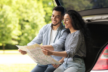 Happy young lovers sitting on auto trunk and watching road map, copy space. Handsome arab guy bonding with his beautiful girlfriend, having break while car trip, looking for destination to go