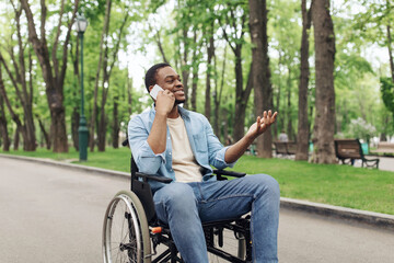 Young black guy in wheelchair having phone conversation at city park in summer. African American man with impairment using smartphone, talking to friend, enjoying remote communication outdoors