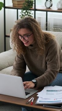 Vertical video of a happy woman working on a laptop at home. She smiles while typing and then thoughtfully continues her work on the couch in the living room.