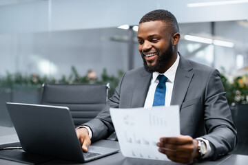 A man in a business suit sits at a desk with a laptop open and looks at documents. He smiles while analyzing data in a modern office space filled with plants.