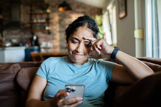 Adult woman worried while reading smartphone on sofa at home