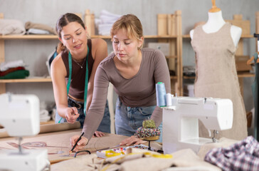 Two dedicated young female dress designers work side by side in creative fashion studio, developing new patterns, drawing and adjusting layout for future garment on paper