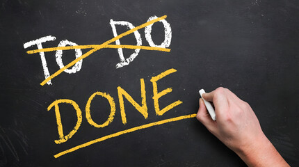 A hand writes the word DONE in yellow chalk on a blackboard as TO DO is crossed out conveying successful task completion and accomplishment