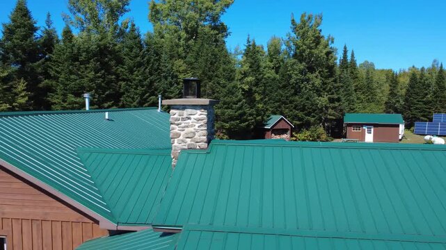 A rock and mortar chimney in the center of a green metallic roof in a forest camp with solar panels under a cloudless blue sky.