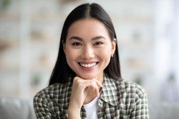 Fototapeta premium Closeup potrait of attractive long-haired asian woman in cozy outfit chilling at home, smiling at camera, copy space. Home interior, millennials lifestyle, relaxation and leisure concept