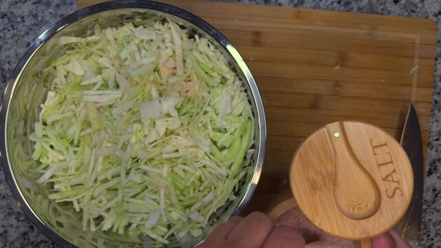 top view of hands adding pink himalayan salt to shredded cabbage in a bowl