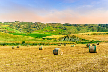 beautiful golden valley with green hills and fields with hay stacks with mountains on background and scenic colorful cloudy sunset on background of landscape