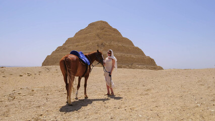 Woman enjoying a vacation in Egypt, standing beside a horse near ancient pyramids in the desert under the bright sun.