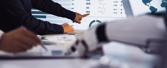 Professionals engage in data analysis, utilizing a robotic hand and digital screen to navigate financial trends and insights during a collaborative workplace discussion.Noogenesis