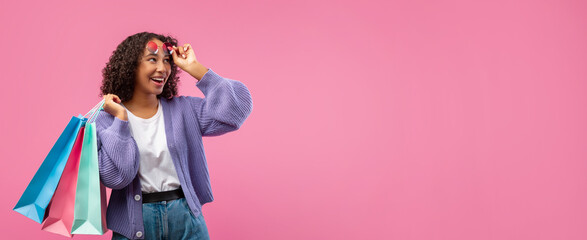 A young woman holds shopping bags while smiling and looking to the side. She wears a light purple...