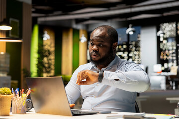 Black office employee checking the time on wristwatch and working late, trying to hurry up and finish his tasks at midnight. Busy fatigued businessman doing overtime alone at the office.