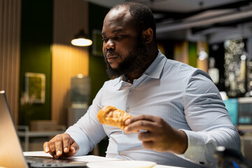 Black man analyst does emotional compulsive overeating due to workload at workstation, eating junk food and pastry as an unhealthy lifestyle. Emotional stress eating disorder at night.