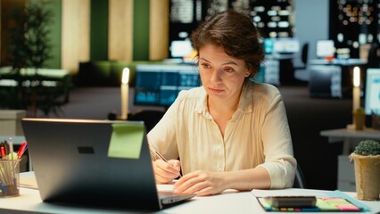 Corporate employee focused on writing a detailed report under a desk lamp. Summarizing updates, organizing the agenda and drafting insights for the corporation management plans. Camera B.