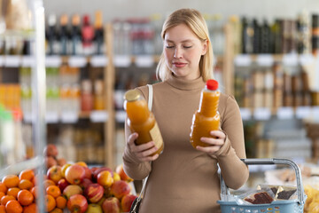 Girl choose and pick up peach juice in store. Customer consider and make choice, compare some varieties products. Client hesitate before buying, choose fruit juice, read labels