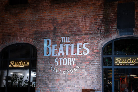 The Beatles Story museum facade at Liverpool Albert Dock is lit at night. Silver lettering shines as Rudy's Pizza Napoletana neon glows through arches, with cool lighting.