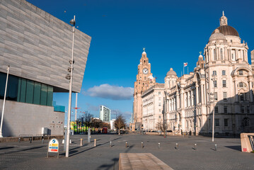 Winter sun illuminates Liverpool Pier Head. Royal Liver Building and Port of Liverpool Building stand near Museum of Liverpool. Sparse pedestrians cast long shadows.