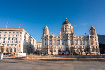 Obraz premium Street level view in Liverpool shows Port of Liverpool Building and Union Jack flags as a red double decker bus passes the waterfront promenade in winter light.