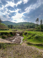 Wide view of a small river crossing with a stone bridge in a fertile green landscape under a dramatic sky in Rwanda.