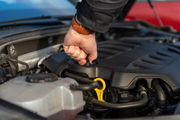 Hand with leather watch strap opening oil filler cap on black engine cover with yellow dipstick and coolant tank visible in car engine bay. Concept of DIY oil change, vehicle maintenance and car care.
