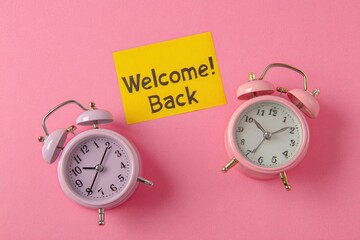 A cheerful and inviting still life composition features two classic pink alarm clocks symmetrically placed on a vibrant, textured pink background. Nestled between the timepieces is a bright yellow sti