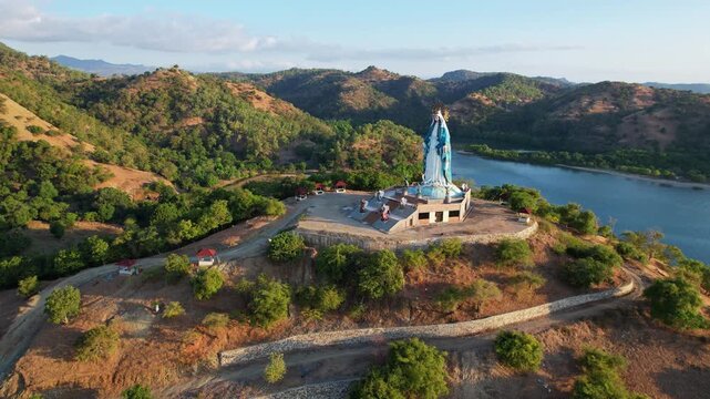 Aerial drone footage of Maria tall colorful statue throning on a dry hill, with small mountains landscape in the background, orange sunset time, in Teluk Gurita, Belu Regency, Timor island, Indonesia
