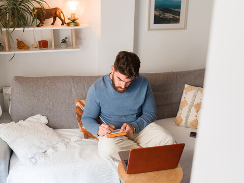 Man writing notes while working ta home with laptop 