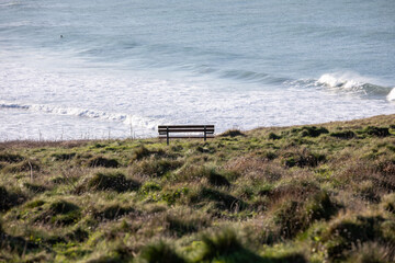 Empty wooden memorial bench on a grassy cliff edge at Pentire Head, Cornwall, overlooking the blue Atlantic Ocean on a clear day.