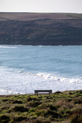 Empty wooden memorial bench on a grassy cliff edge at Pentire Head, Cornwall, overlooking the blue Atlantic Ocean on a clear day.