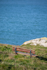 Empty wooden memorial bench on a grassy cliff edge at Pentire Head, Cornwall, overlooking the blue Atlantic Ocean on a clear day.