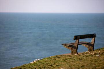 Empty wooden memorial bench on a grassy cliff edge at Pentire Head, Cornwall, overlooking the blue Atlantic Ocean on a clear day.