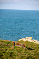 Empty wooden memorial bench on a grassy cliff edge at Pentire Head, Cornwall, overlooking the blue Atlantic Ocean on a clear day.