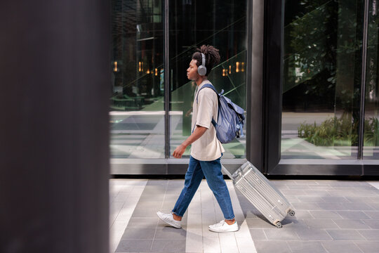 Traveler with headphones and luggage walks past modern building.