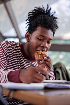 Young man eats muffin while writing in notebook.