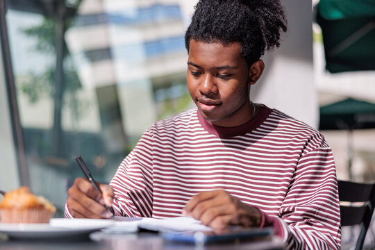Young man writing in notebook at outdoor cafe.