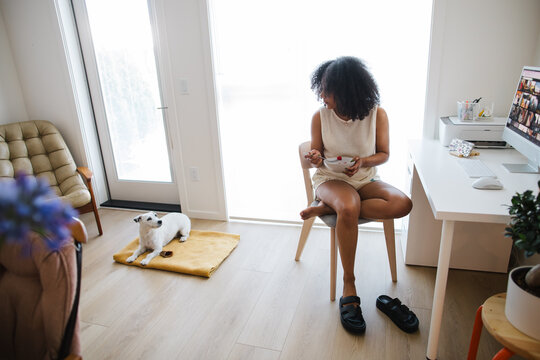 Candid home office scene: woman eating at desk with dog nearby i