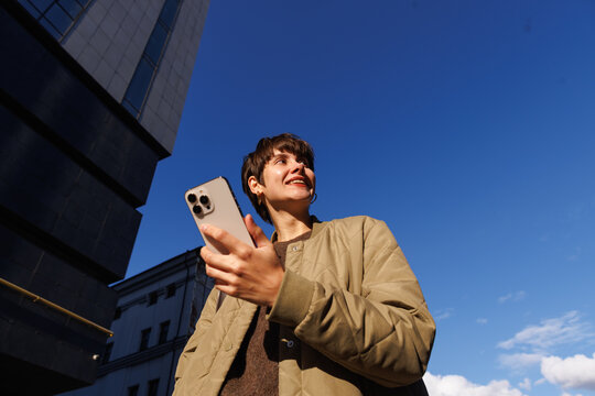 Smiling woman holding smartphone in city sunlight against blue sky