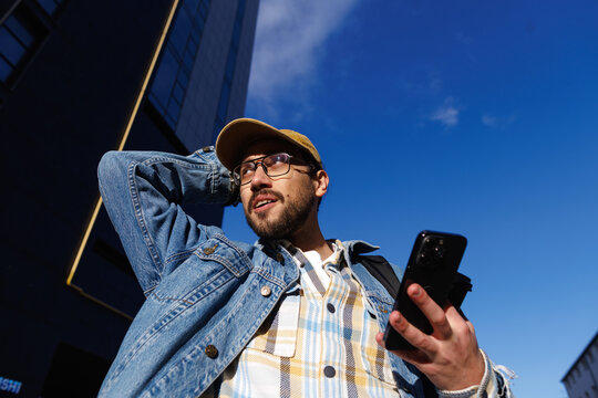 Casual man with smartphone looking up outdoors against blue sky