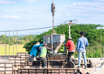 Construction workers manually guiding a large crane-suspended concrete bucket to pour wet cement into foundation formwork