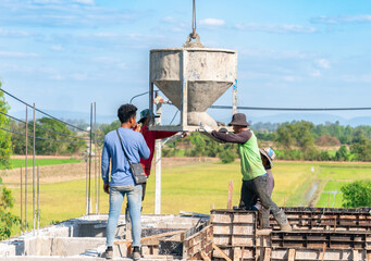 Construction workers manually guiding a large crane-lifted concrete bucket to pour wet cement into foundation formwork