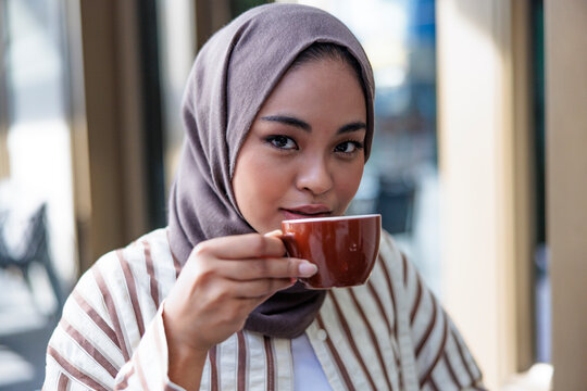 Woman in hijab sips coffee, looking at the viewer.
