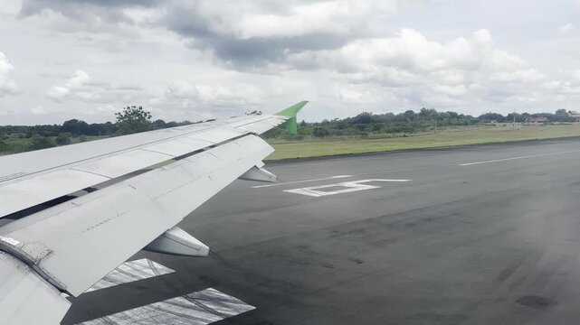 Passenger view from airplane window during taxiing on the runway before takeoff, featuring the aircraft wing with green winglet against a cloudy sky and airport landscape background.