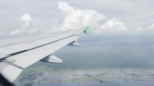 Aerial view of a commercial airplane wing with a green tip flying over a lush tropical forest landscape and river under a cloudy sky.