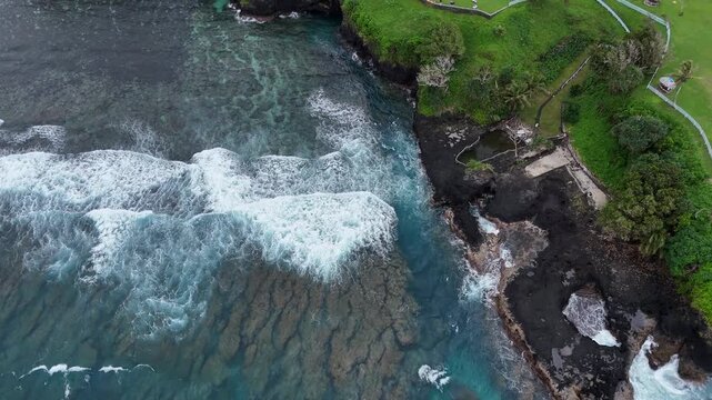 Dramatic aerial drone shot of powerful Pacific waves crashing against the rugged volcanic shoreline of Samoa, creating beautiful patterns of white foam and surf.