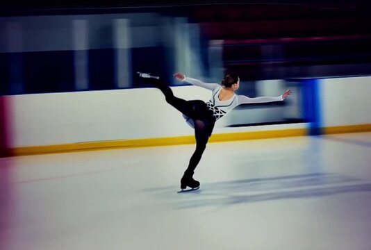 Female skater in white and black outfit holds a high arabesque as the camera pans. Indoor arena, white boards, yellow kickplate, red exit lights, cool lighting.