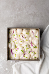 overhead view of slices of onion and scallion focaccia dough in a square baking pan, top view of airy bubbly onion focaccia dough proofing on a cement countertop