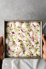 overhead view of slices of onion and scallion focaccia dough in a square baking pan, top view of airy bubbly onion focaccia dough proofing on a cement countertop