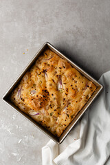 overhead view of onion and scallion focaccia in a baking tin, top view of airy bubbly onion focaccia on a cement countertop	